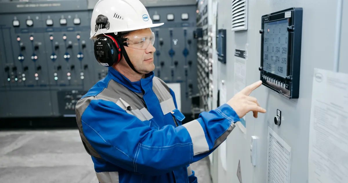 Técnico con casco blanco y orejeras señala la lectura de un panel de control digital en la sala de tableros de una planta industrial