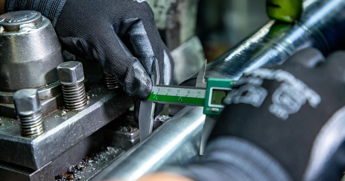 Manos con guante negro miden con un calibrador digital verde una barra metálica sujeta junto a una pieza mecanizada con virutas