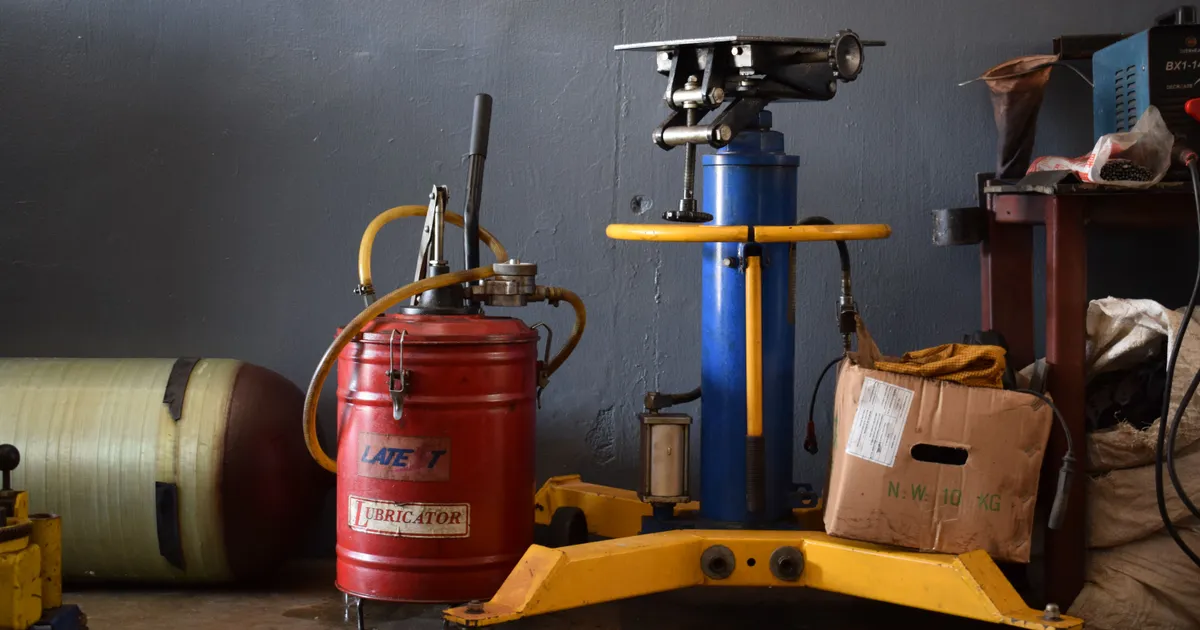 Red lubricator drum with hose and hand pump standing next to a blue hydraulic jack in a workshop