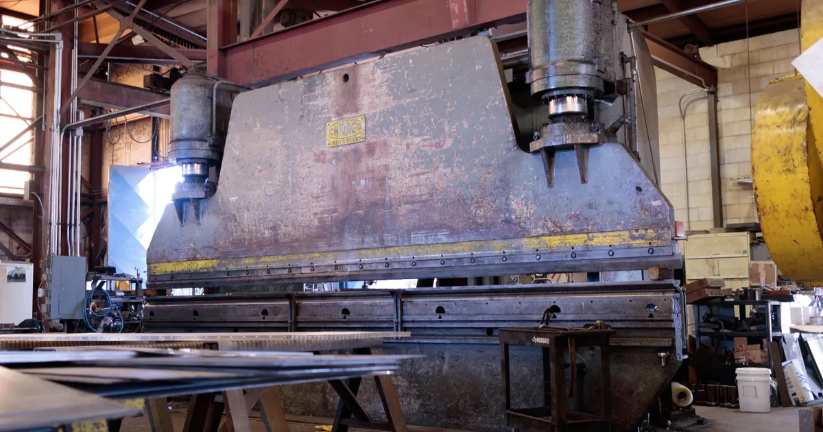 Massive steel press filling a factory bay, seen from floor level with its heavy ram, bed and two hydraulic cylinders overhead