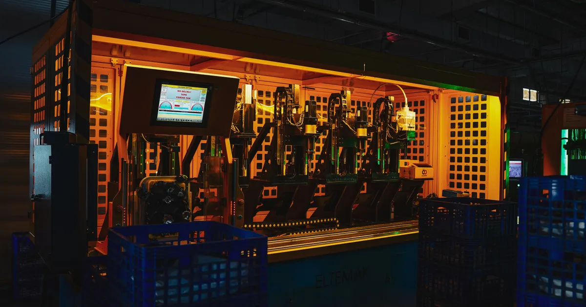 Guarded automated production cell lit in orange, with a control screen, a row of machine stations and blue stacking crates in the foreground